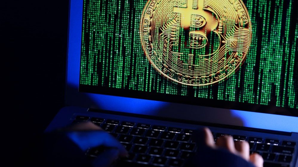 Cyber criminals in a dark room using a monitor with the Bitcoin logo shown