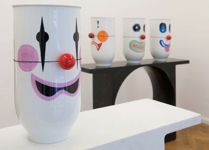 Different expression of clown faces on charpin vases. Three of the Vases are on a black marble stand with arch center. One is on a white stand. All photographed against a white background and wood floor