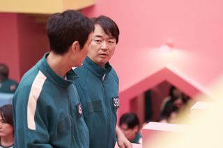 A man (Lee Seo-hwan) stands next to another man in a bright pink room, as they wear matching green tracksuits, in 'Squid Game' season 2.