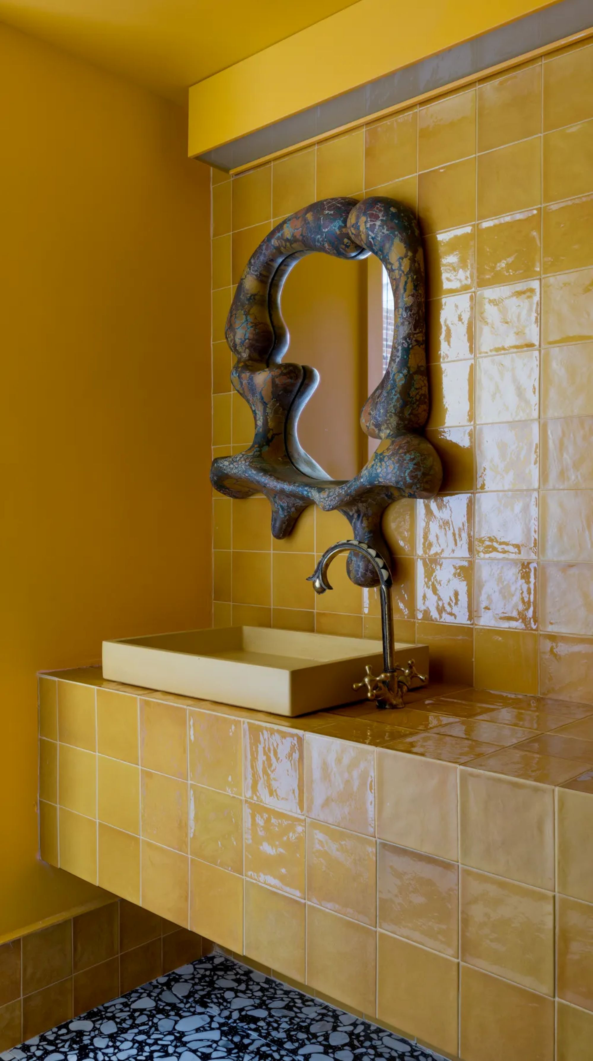 Image of a yellow bathroom with a sink and backsplash made out of large yellow tile. The floor is a black and white terrazzo material. There is a blueish-gray mirror over the sink in an abstract shape.
