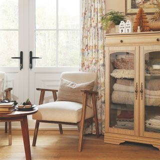 Glass-fronted cabinet filled with blankets in a living room next to an accent chair and table