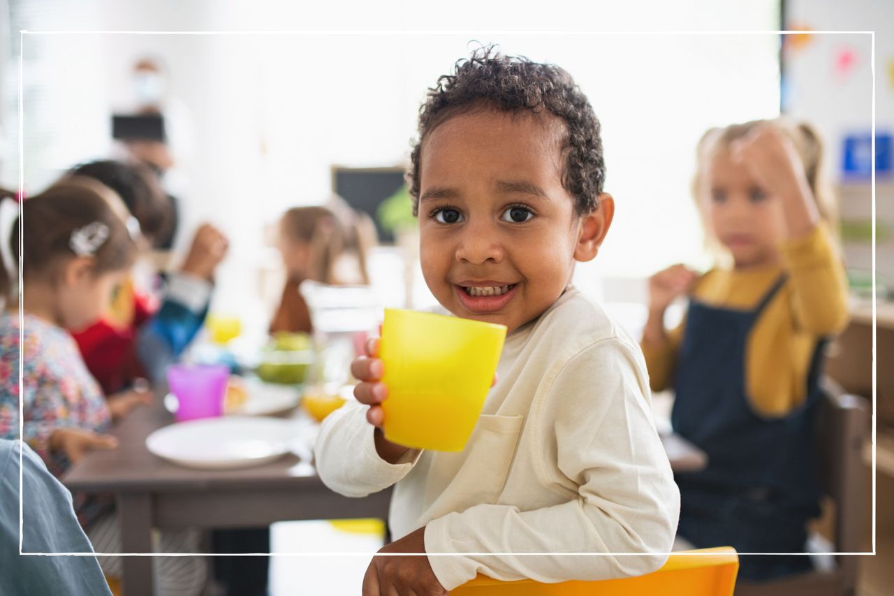 Young child sat at a table at nursery. He turns and looks at the camera with a beaker in his hands