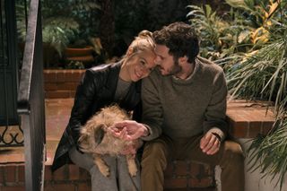 A woman (Kristen Bell as Joanne) holds a rescue dog and rests her head on a man's (Adam Brody as Noah) shoulder as they sit on brick outdoor steps, in episode 103 of Nobody Wants This.