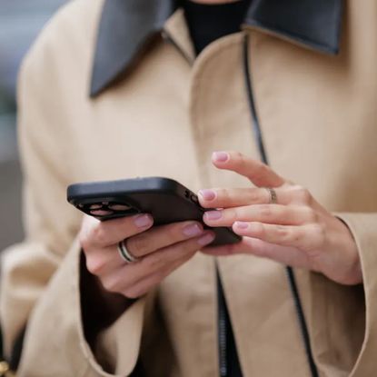 woman wearing beige trench and holding mobile phone with milky pink manicure 