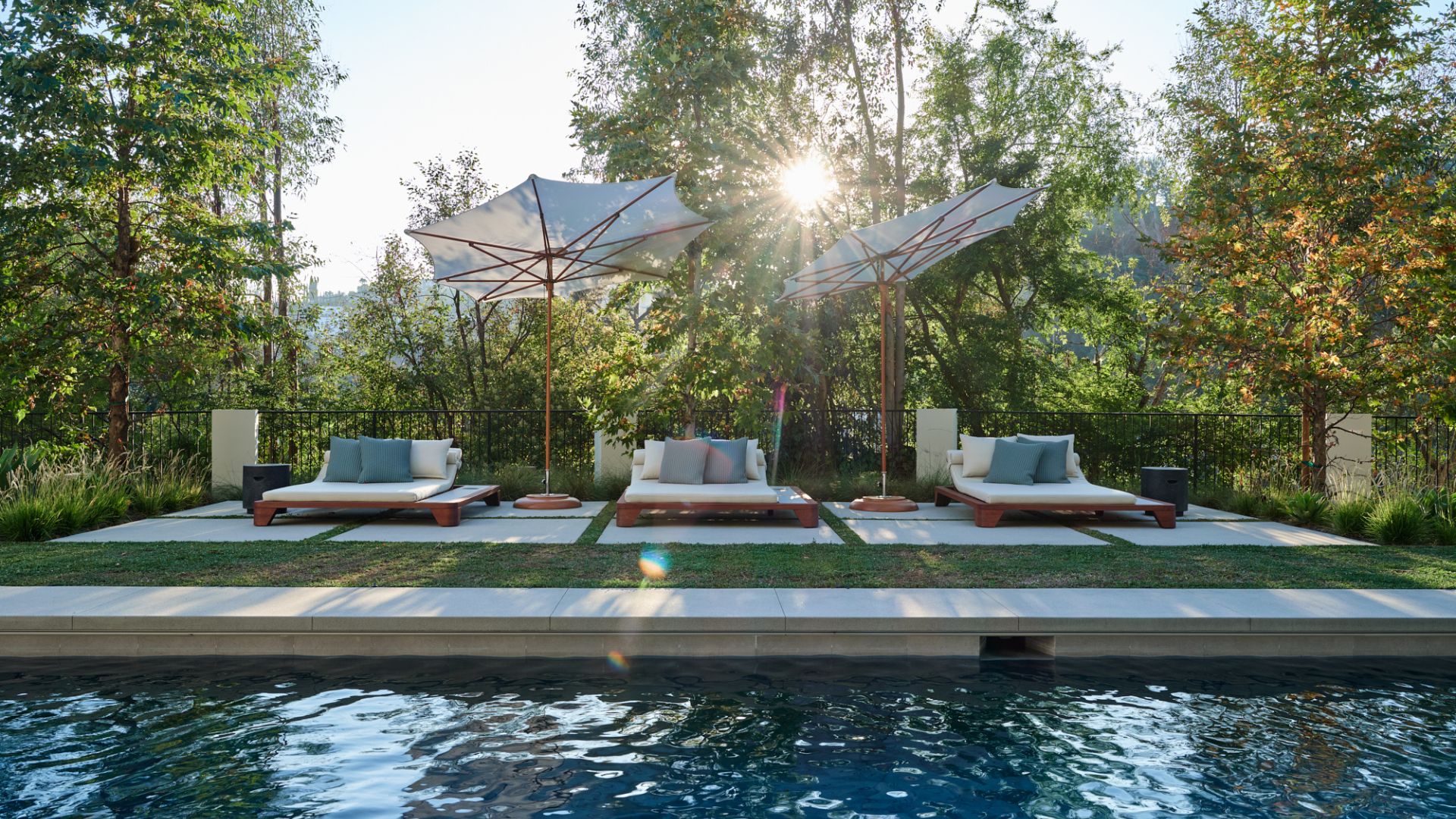 A backyard with a pool and three sun loungers under two large parasols