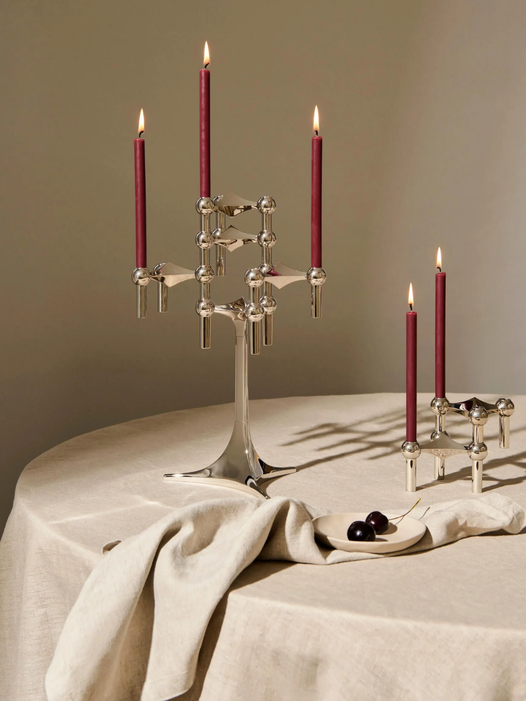 A table with a beige table cloth, a bowl of cherries and red dinner candles on silver stacked holders