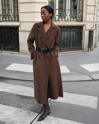 Paris woman wears a brown Jenni Kayne wool coat with a black waist belt, sheer tights, and black kitten heels.