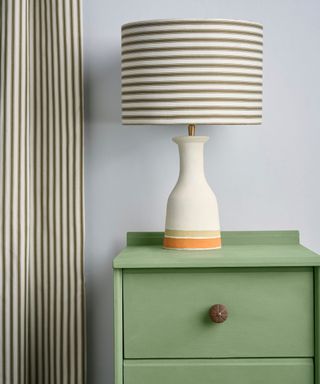 A white lamp with a stripey lampshade on a green chest of drawers