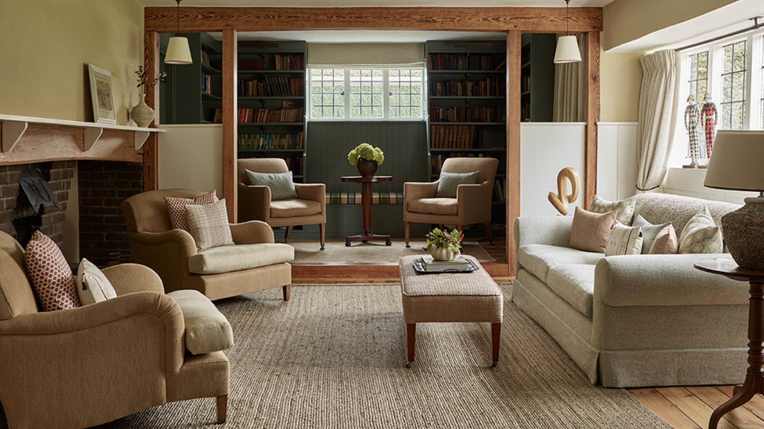 a cozy cottage living room in hampstead with a neutral rug, inglenook fireplace, a beamed nook area with dark green bookcases and layered furniture