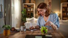 Young woman carefully managing her home finances, putting coins in a piggy bank and taking notes on expenses and savings in her living room