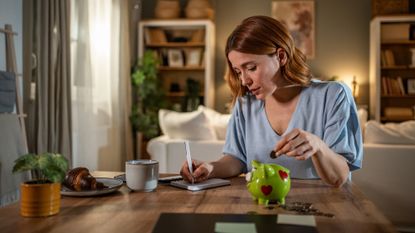 Young woman carefully managing her home finances, putting coins in a piggy bank and taking notes on expenses and savings in her living room