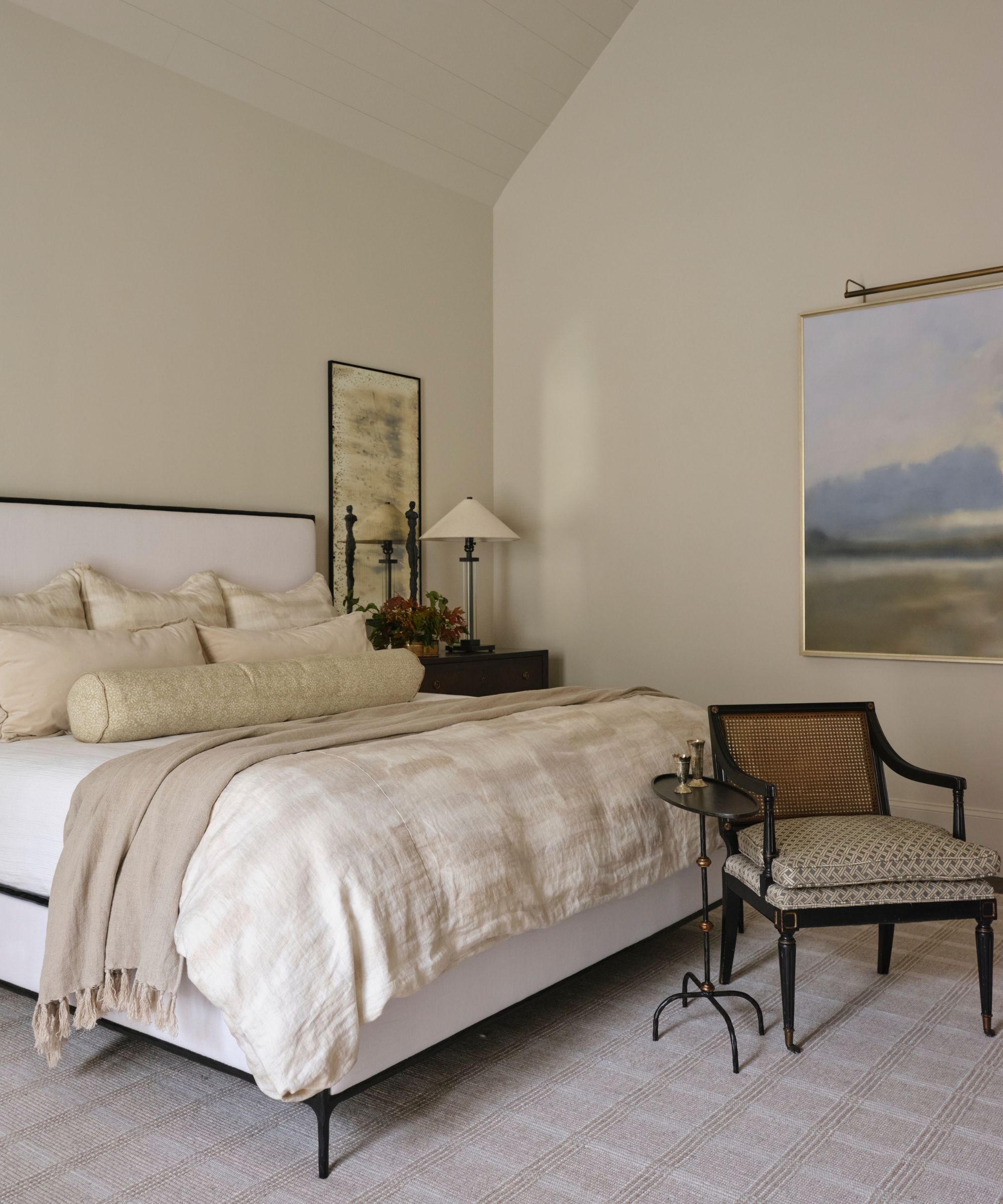a neutral primary bedroom with white and iron bed, neutral linens, checked carpet and antique table and chair