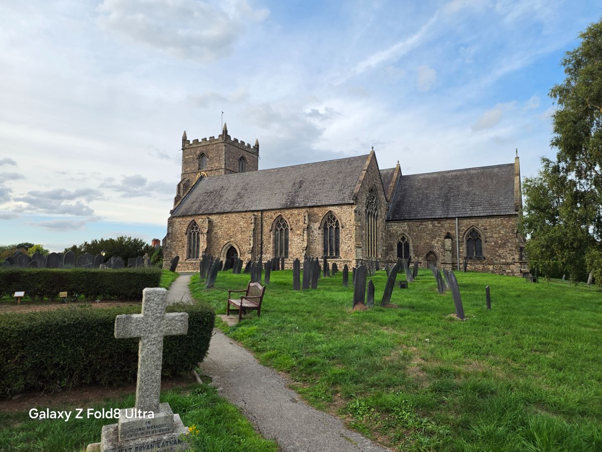 Historic stone church with Gothic windows, gravestones and a large stone cross in the foreground.