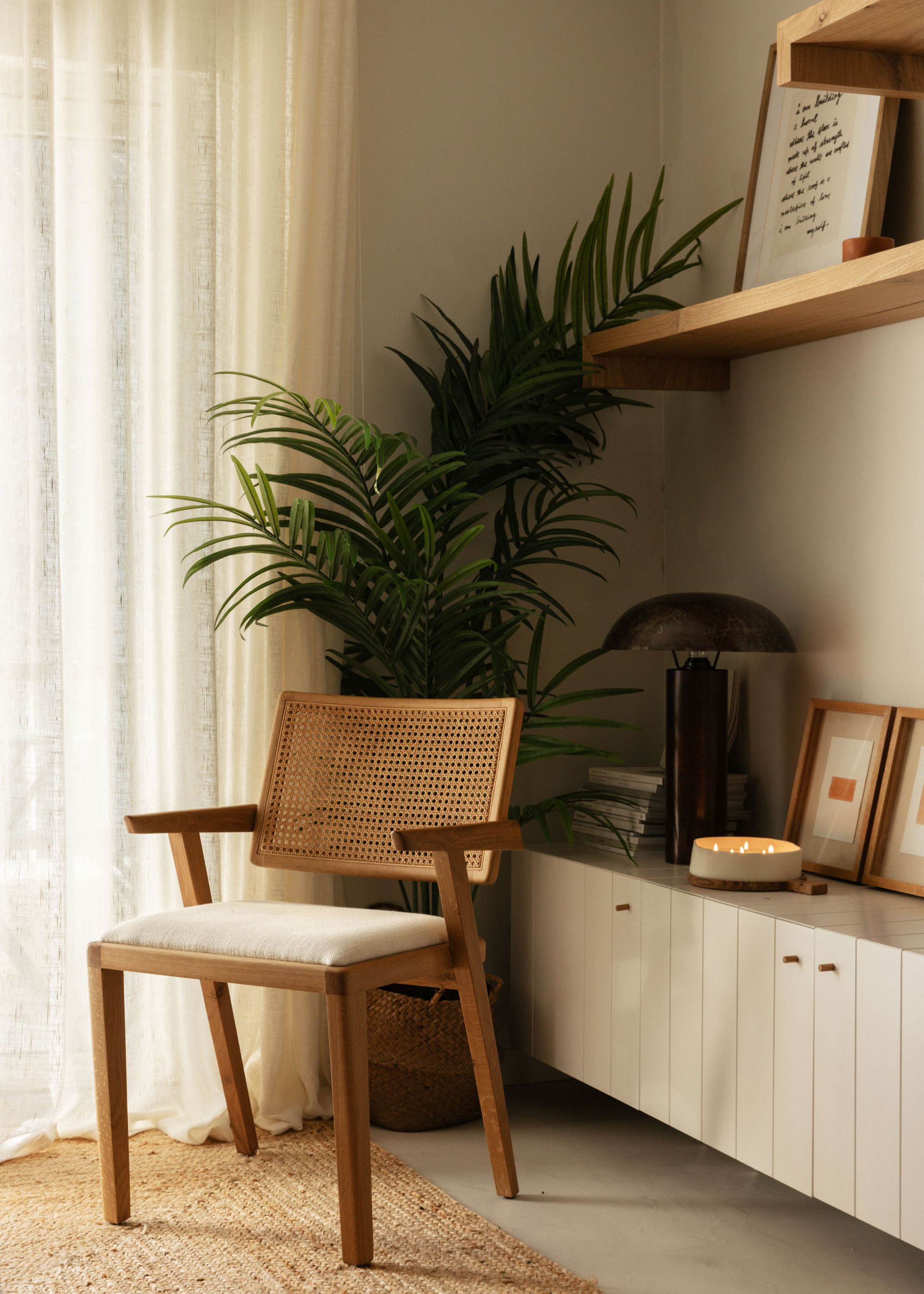 A calm living room corner with a green plant, a rattan chair, a woven natural rug, a floating white sideboard holding a multi-wick candle, a black lamp, framed prints and a stack of books