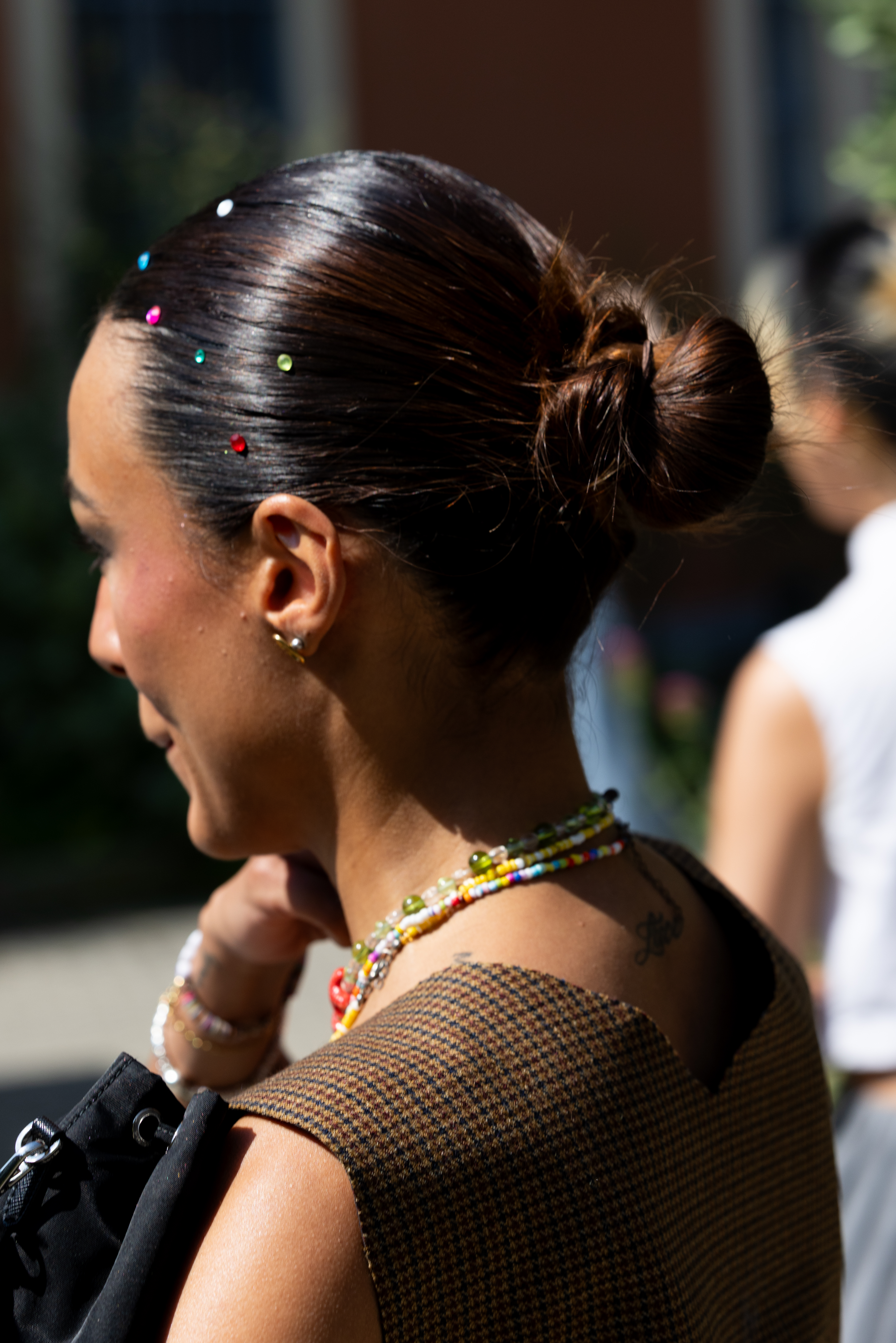 A woman with colored gem stones in her slicked back bun hairstyle. She's also wearing beaded necklaces and a brown top.