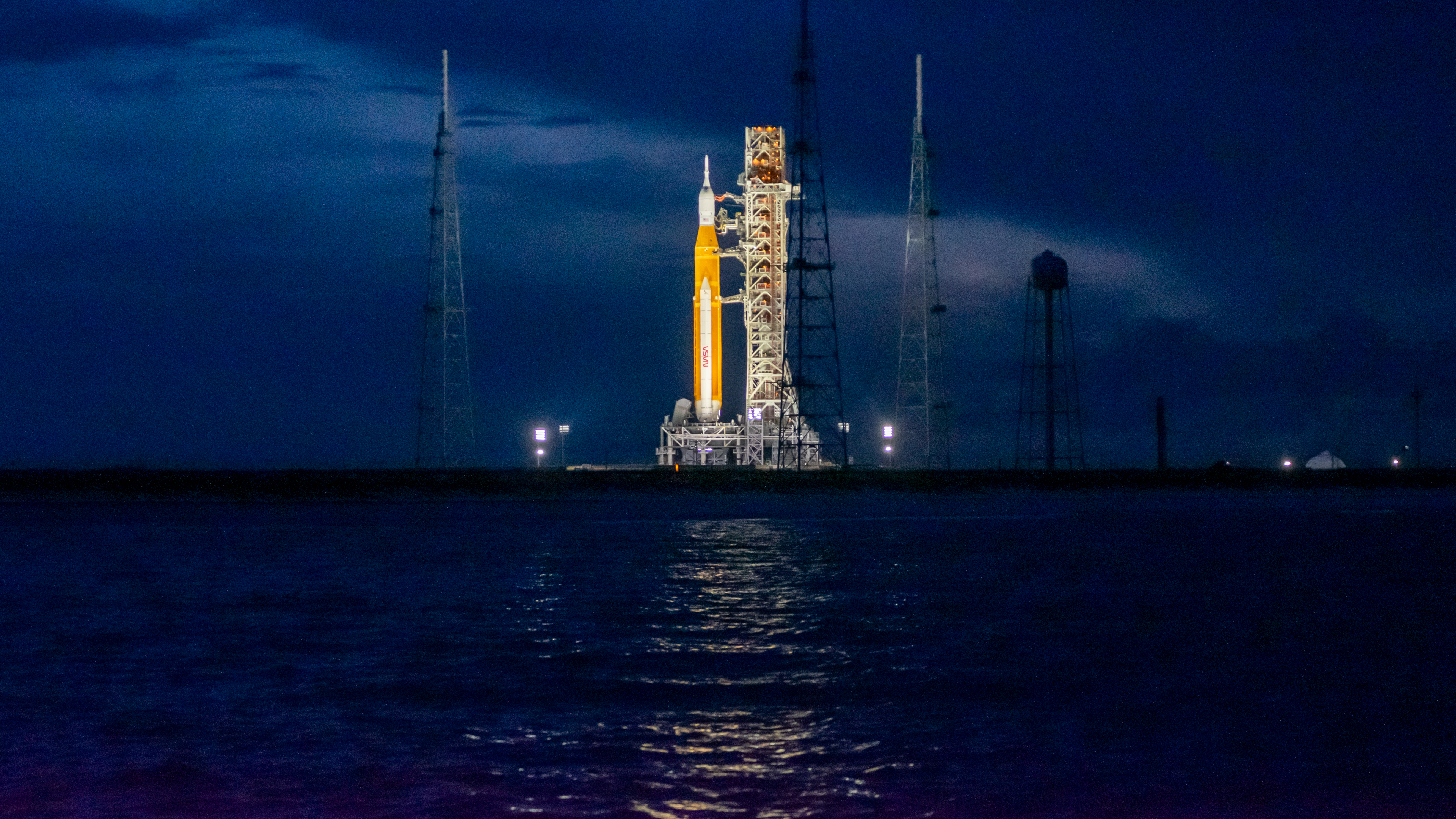 a bright orange rocket stands next to a grey tower illuminated amid a dark blue background sky and rippling sea in the foreground.