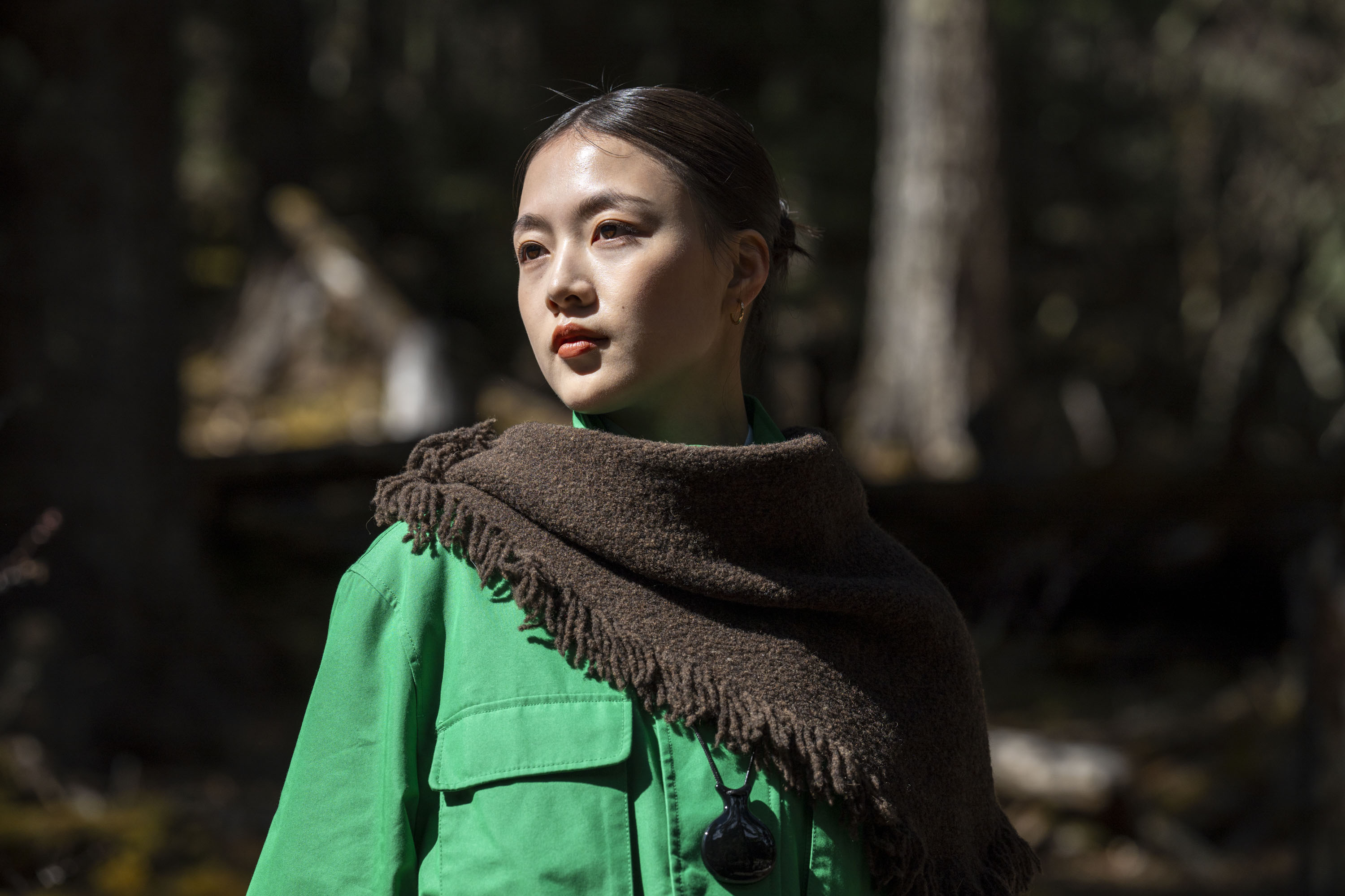 Portrait of a woman in a shawl and green jacket in a woodland, there's direct sunlight on her