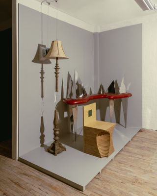 Vintage design objects including a standing lamp, a futuristic mirror and resin desk, and a wooden chair are captured in a film photograph as they sit in a gallery.