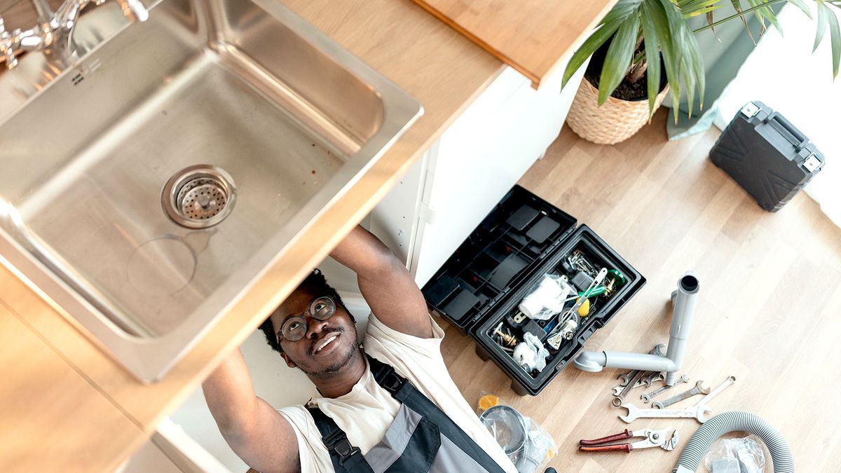 Man under stainless steel sink fitting pipework with tools next to him on wooden floor