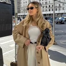 style influencer Fredrika Ekerot poses on a NYC sidewalk wearing black oval sunglasses, a white tank top, oversized tan trench coat, black belt with a silver buckle, a black woven bag, and beige pants