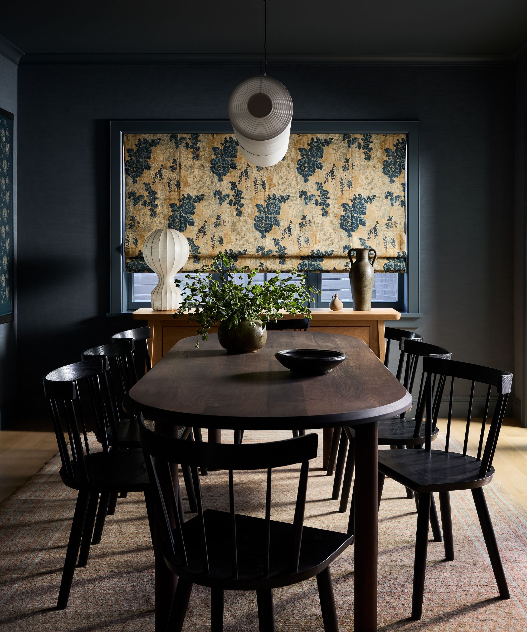 a dark dining room with wooden tables and chairs, vintage rug, floral window blind and a large sculptural chandelier