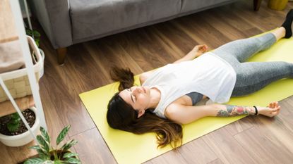 woman lying on her back on a yellow mat in a living room setting doing a relaxation practice. 