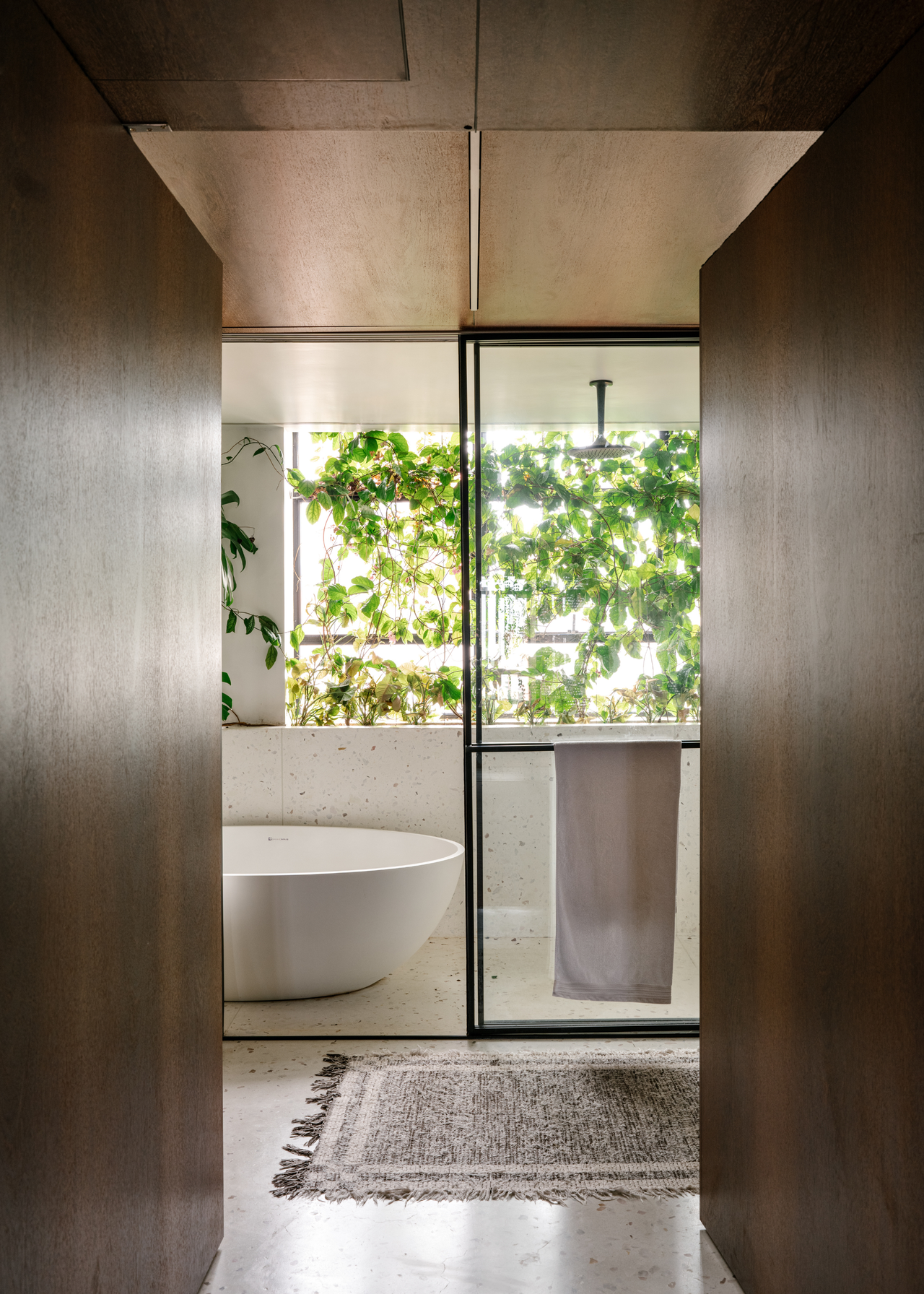 A set of wooden double doors leading into a bathroom with a shower cubicle and standing tub witha screen of foliage