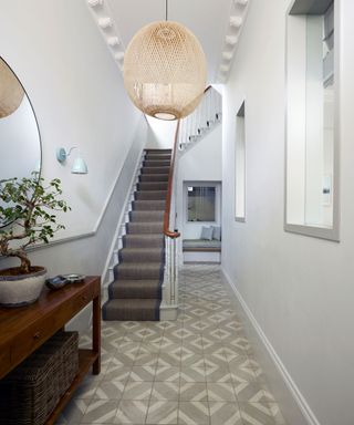 Hallway with grey and white tiled floor, staircase with stair runner and large woven lampshade hanging below a coved ceiling.