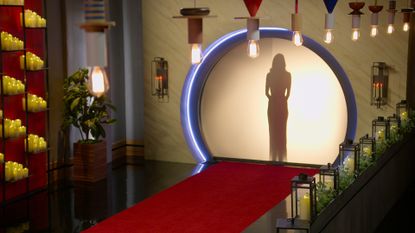 A woman stands behind a circular backlit screen, which opens into a chamber decorated with flowers, Edison bulb lights, candles, and a red carpet, in &#039;Love is Blind&#039;