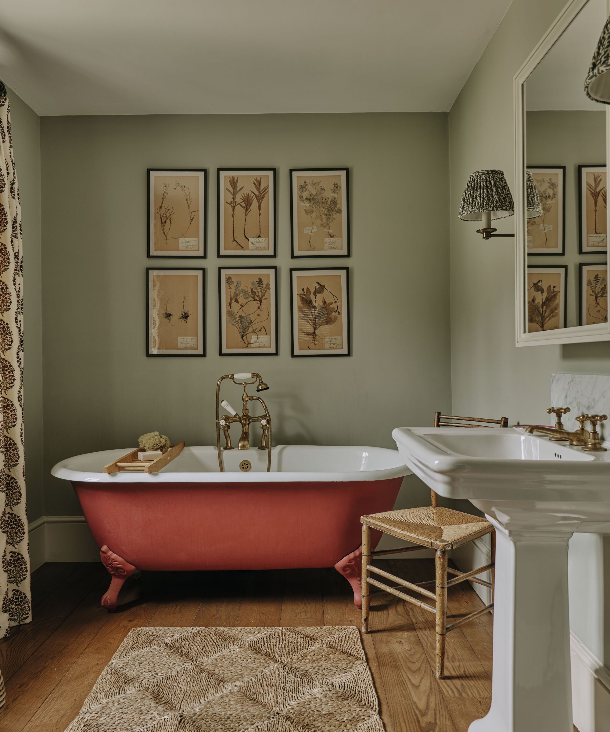 Traditional bathroom with a terracotta-red clawfoot tub, brass fixtures, a pedestal sink, and framed botanical prints on sage green walls