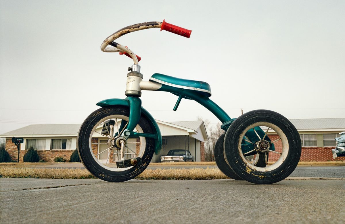 A color photograph of a weathered green child's tricycle parked on a driveway in front of a suburban house.