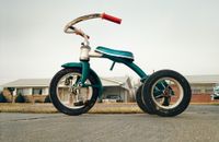 A colour photograph of a weathered green child's tricycle parked on a driveway in front of a suburban house.
