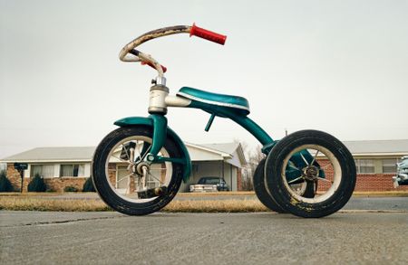 A colour photograph of a weathered green child's tricycle parked on a driveway in front of a suburban house.