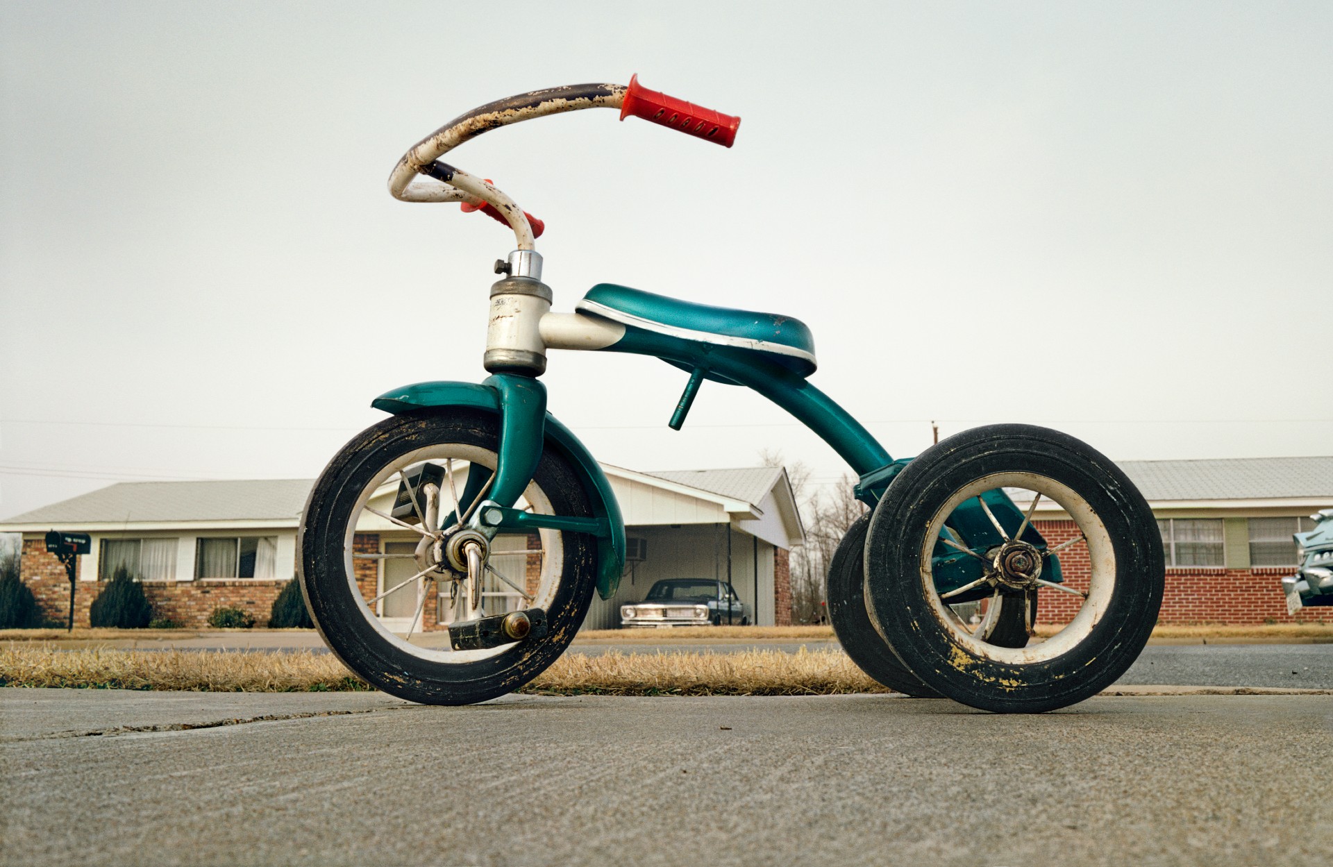 A color photograph of a weathered green child's tricycle parked on a driveway in front of a suburban house.