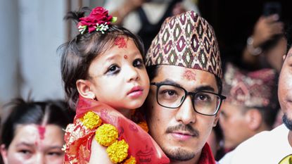 Nepal's appointed Royal Living Goddess, Aryatara Shakya, dressed inn red with a symbolic 'third eye' painted on her forehead, held by an adult man