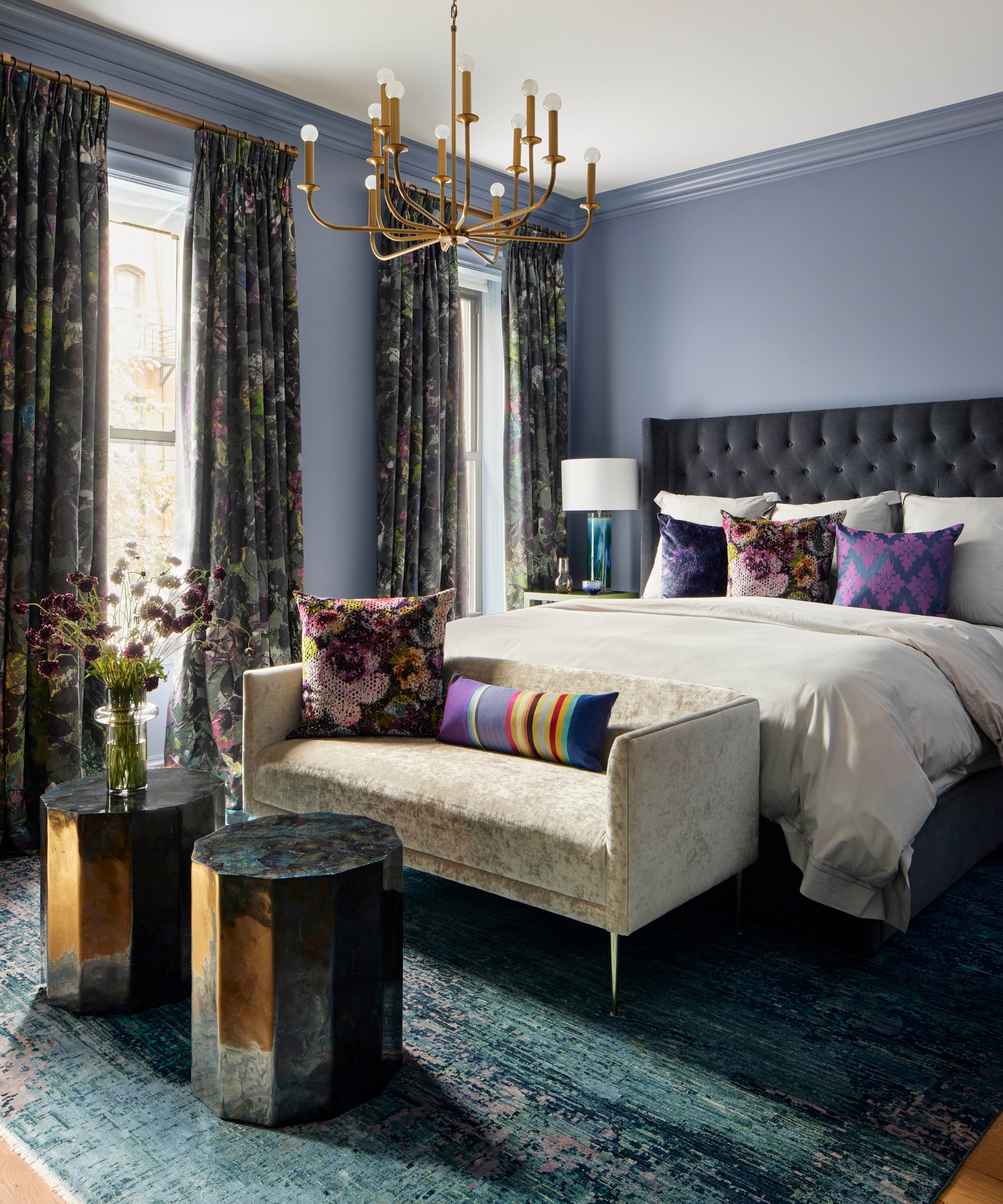 Bedroom with smoky violet-blue walls, floral drapes, brass chandelier, upholstered bed, and jewel-toned accent cushions.