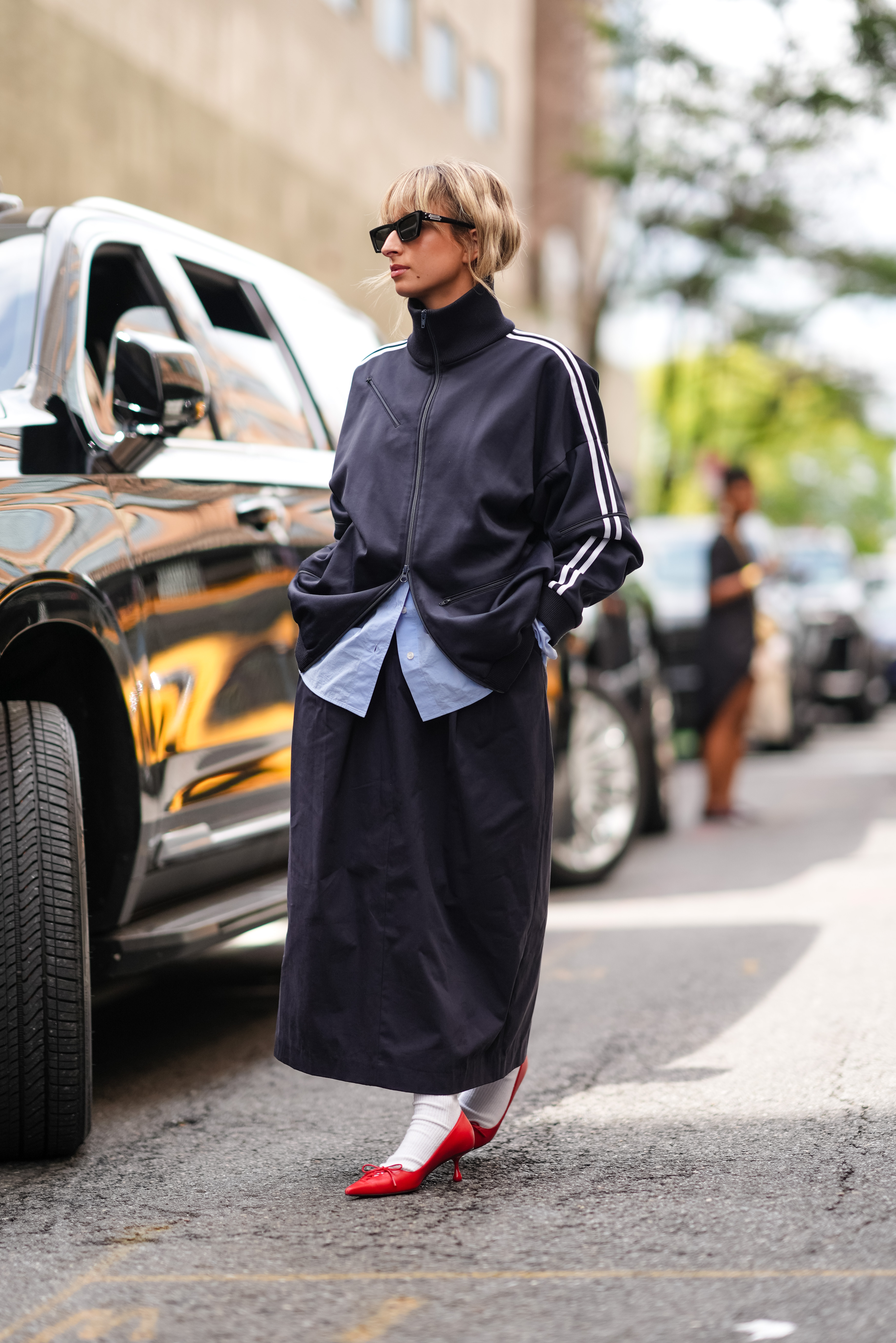 NEW YORK, NEW YORK - SEPTEMBER 07: A guest wears black sunglasses, black high track suit jacket, light blue buttoned up long sleeve shirt, black midi skirt, white socks, shiny red pointed toe ballerina heels, outside Tibi, during the New York Fashion week Spring/Summer 2025 on September 07, 2024 in New York, New York. (Photo by Edward Berthelot/Getty Images)