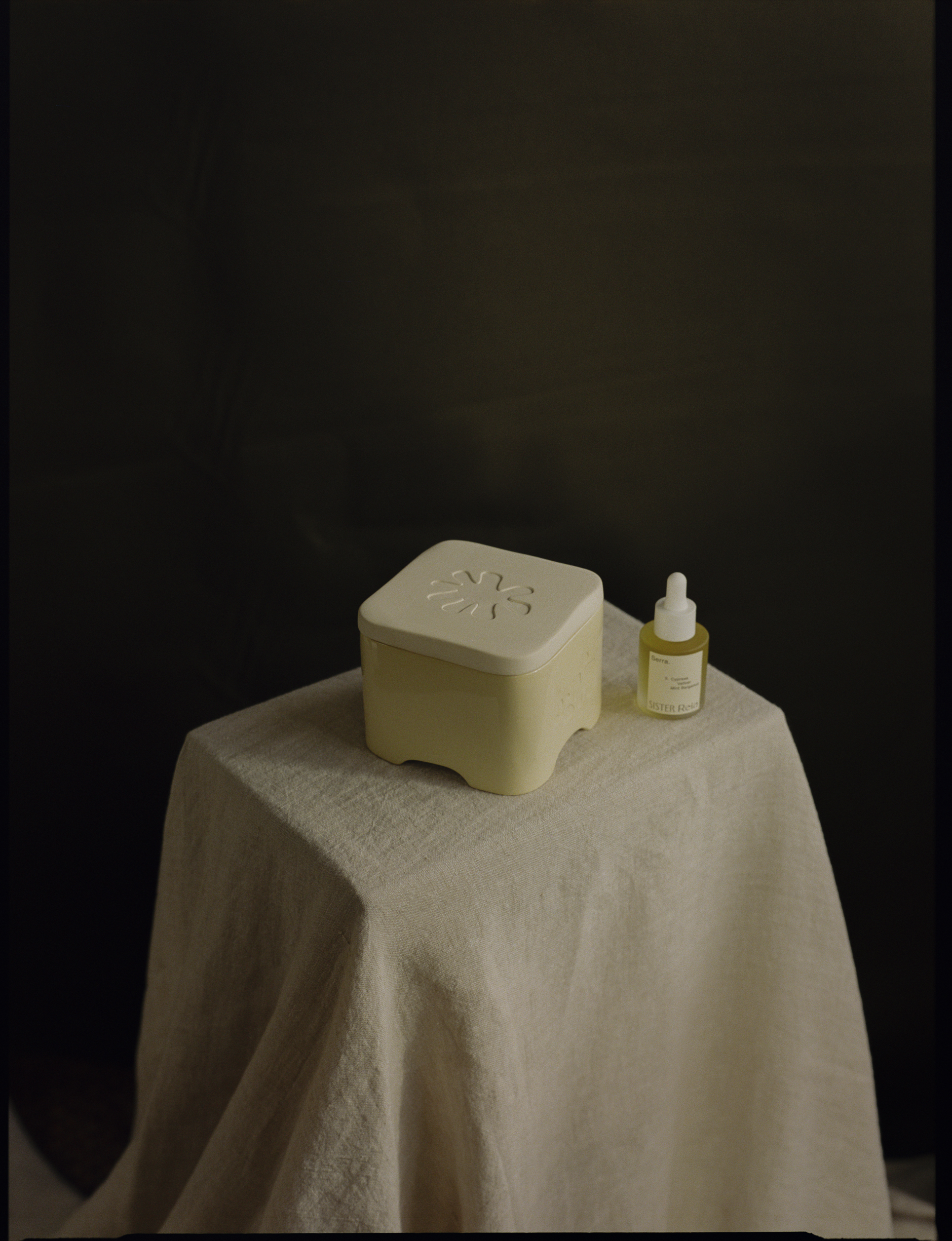 A room lit by a sunset boasts a home fragrance diffuser in the form of a pale yellow box, with a bottle of perfume, a vase of flowers, books, and other collectibles like candles and home accessories.