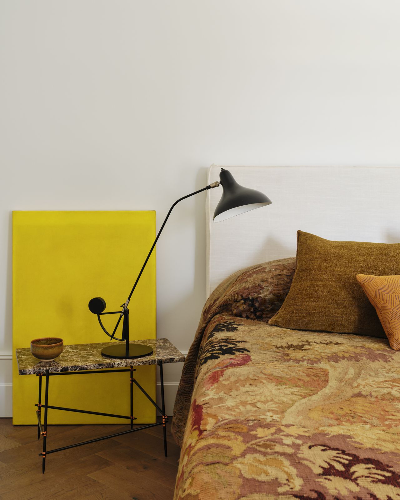 bed covered in tapestry bedcover, marble side table with lamp, and a yellow artwork leaning against the wall