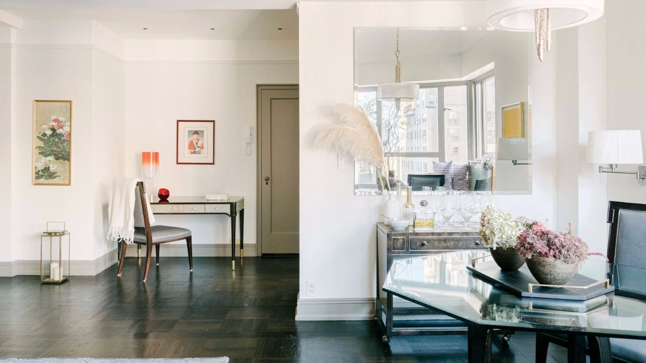 Bright living space with glass tables, chairs, white walls, framed art, a large mirror, and a wooden floor.