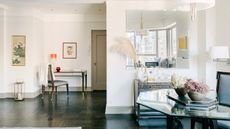 Bright living space with glass tables, chairs, white walls, framed art, a large mirror, and a wooden floor.