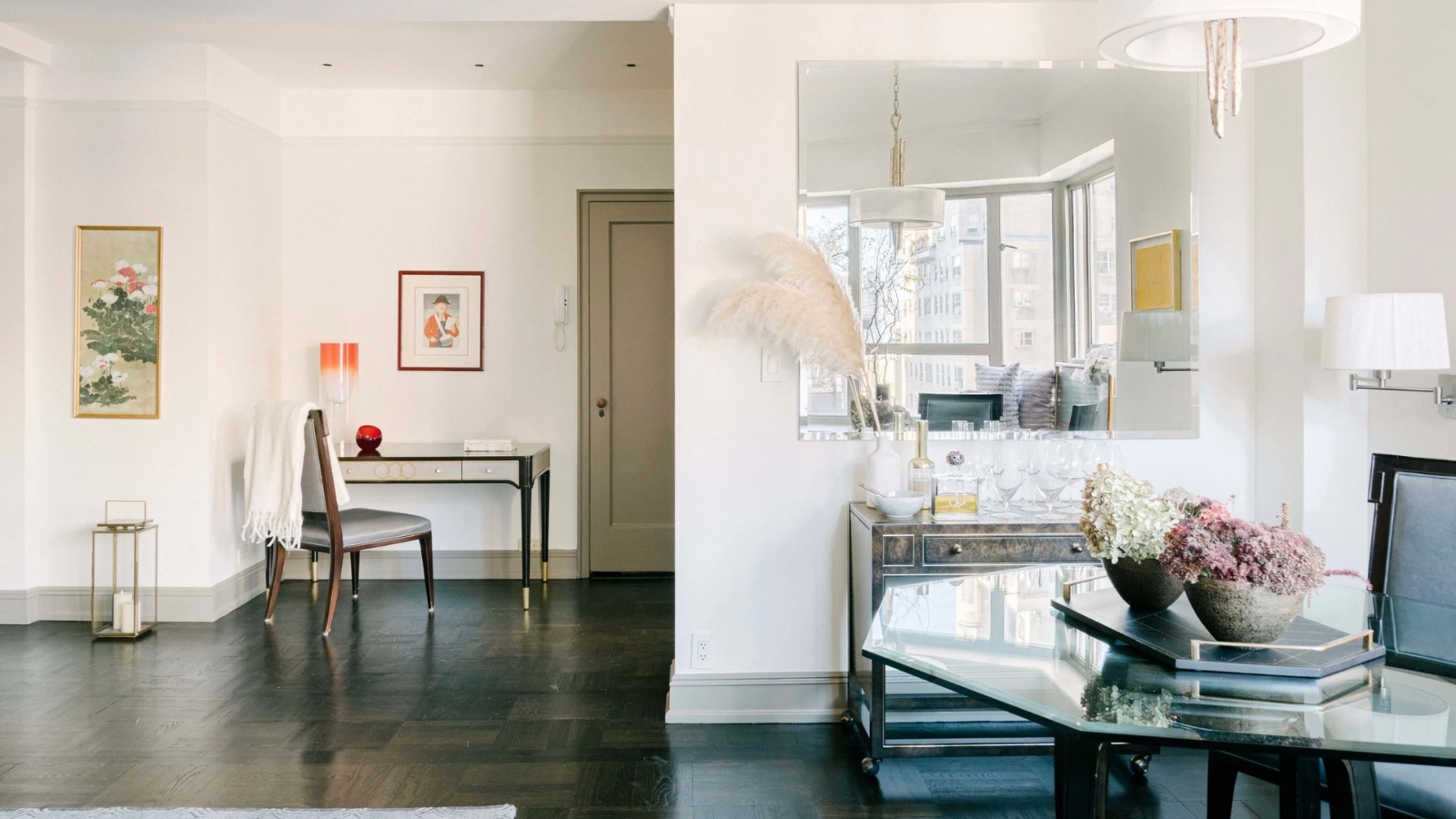 Bright living space with glass tables, chairs, white walls, framed art, a large mirror, and a wooden floor.