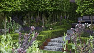 large trees and flowers in a garden within the borders, with a gray garden path lined with small hedges