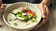 Bowl of food, red and green vergetables, plus mushrooms, in a coconut milk broth. The eater’s hands can be seen holding a spoon over the bowl with the other hand cupping the bowl