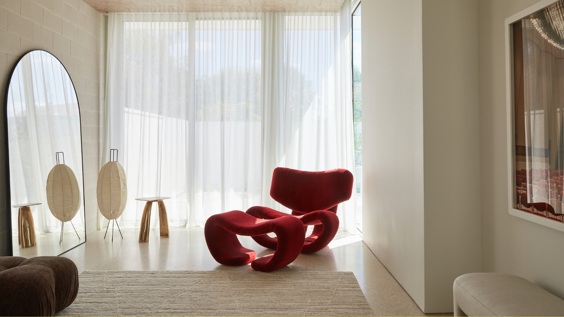 A white living room with an arched mirror, an area rug, a rice paper floor lamp, sheer curtains, a red spine-like chair, and a framed print