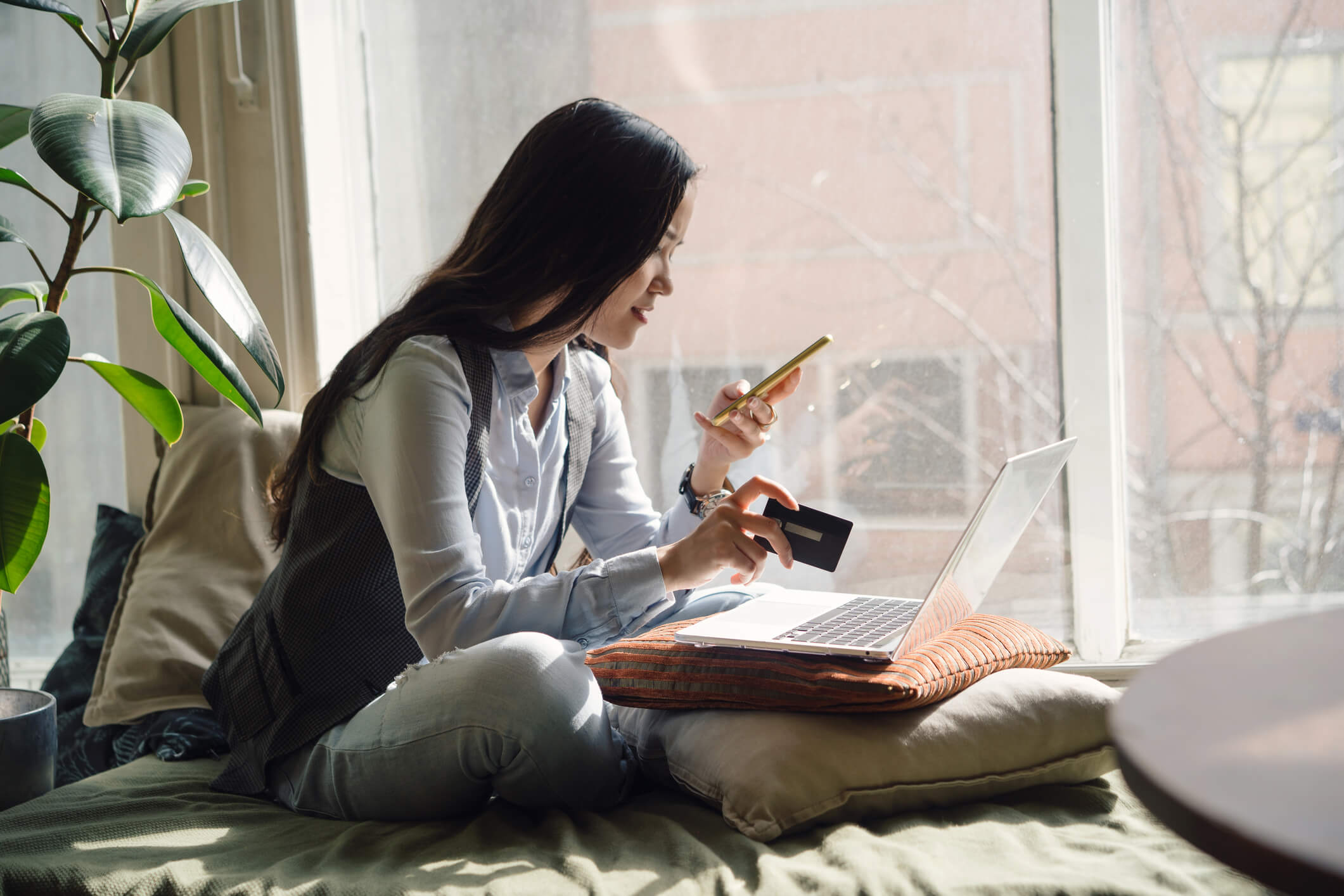 Woman sat down on front of laptop using a phone and holding a credit card