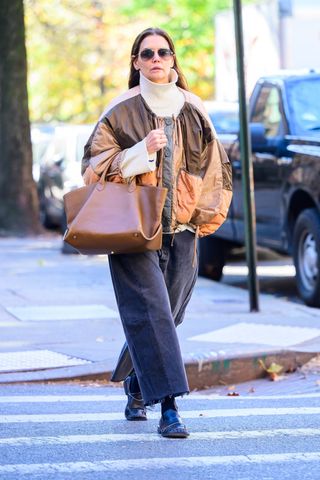 Katie holmes wears a white turtleneck sweater, a brown jacket, tan tote bag, gray jeans, and black square-toe loafers.