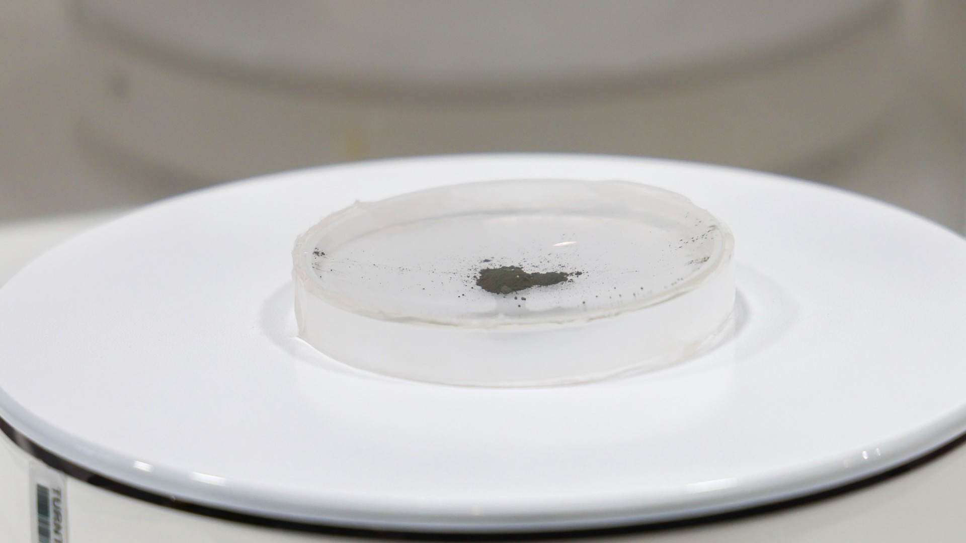 a small sample of grey soil in a glass dish on a white display stand