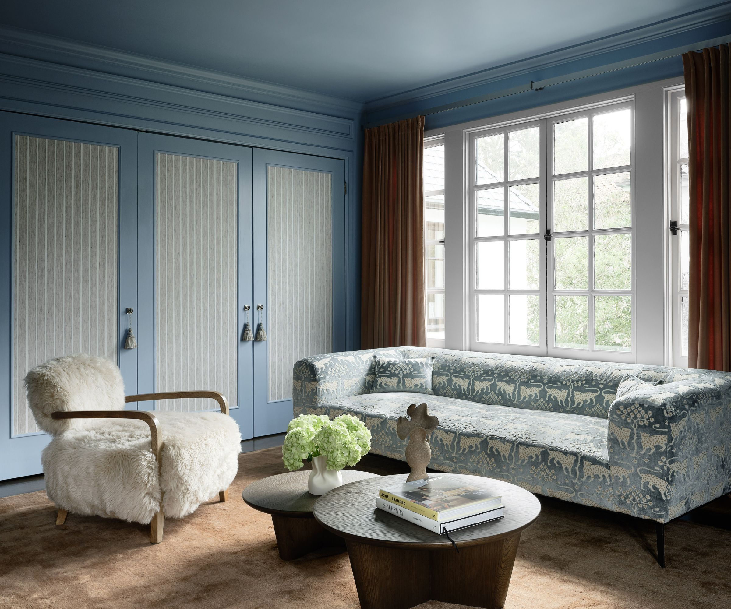 A bedroom color-drenched in a mid-tone blue with a large window with warm brown curtains, a blue and white patterned sofa, a beige rug, two modern circular coffee tables, and a shearling armchair.