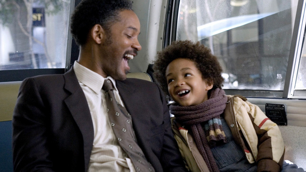 A still from the movie The Pursuit of Happyness in which Will Smith plays Chris Gardner and is sat on a bus laughing with his son Christopher Gardner, Jr.